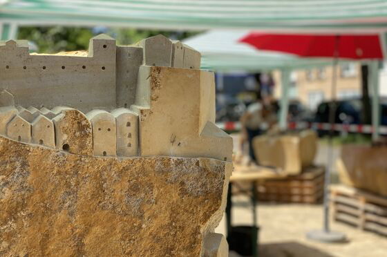Ein Steinklotz, an dem erste Gebäude erkennbar sind, nachdem ihn ein Steinmetz bearbeitet hat. Der Klotz steht unter freiem Himmel. 