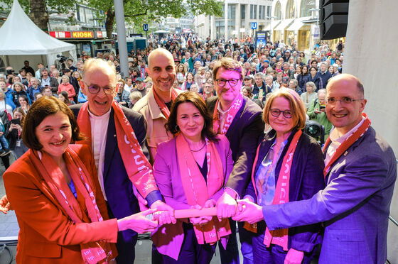 Sieben Personen stehen auf einer Bühne vor Publikum und halten alle gemeinsam einen dicken Stab. 