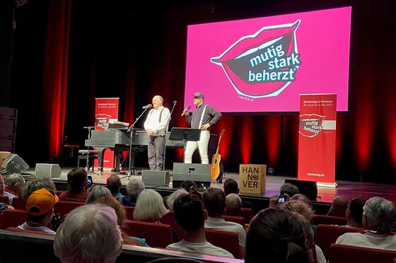 Zwei als Männer lesbare Personen spielen auf einer Theaterbühne vor viel Publikum.