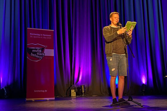 Eine als Mann lesbare Person steht auf einer Bühne und liest aus einem Buch vor.