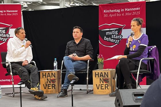 Drei Personen sitzen auf Stühlen auf einem Podium und halten jeweils ein Mikrophon in der Hand. 