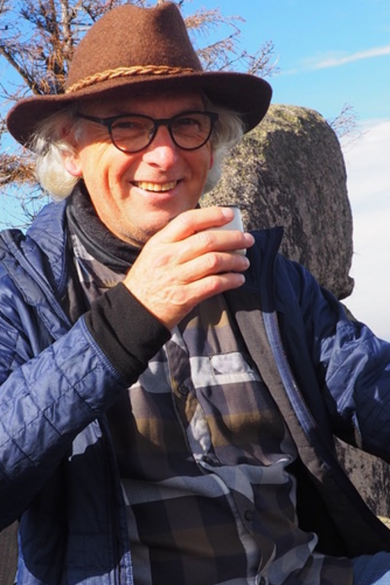 Ein Mann mit Hut sitzt in der Natur. Er lächelt in die Kamera und hält eine Tasse Tee in der Hand.