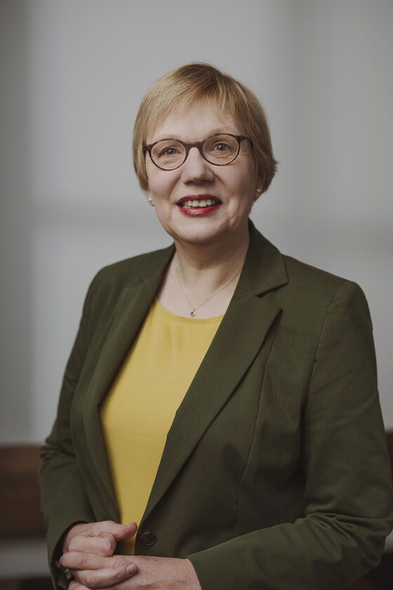 Eine blonde Frau mit kurzen Haaren in gelber Bluse und grünem Jackett mit Brille lächelt.