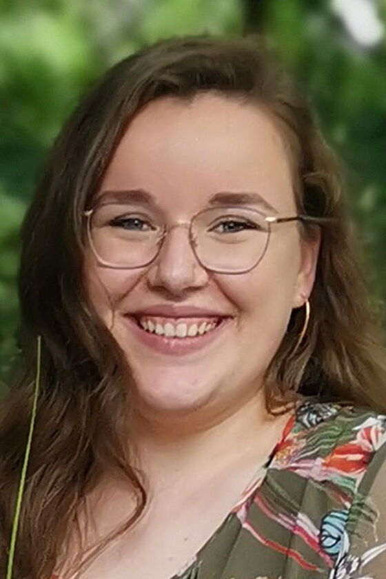 Eine junge Frau mit schulterlangen braunen Haaren und Brille lächelt in die Kamera.