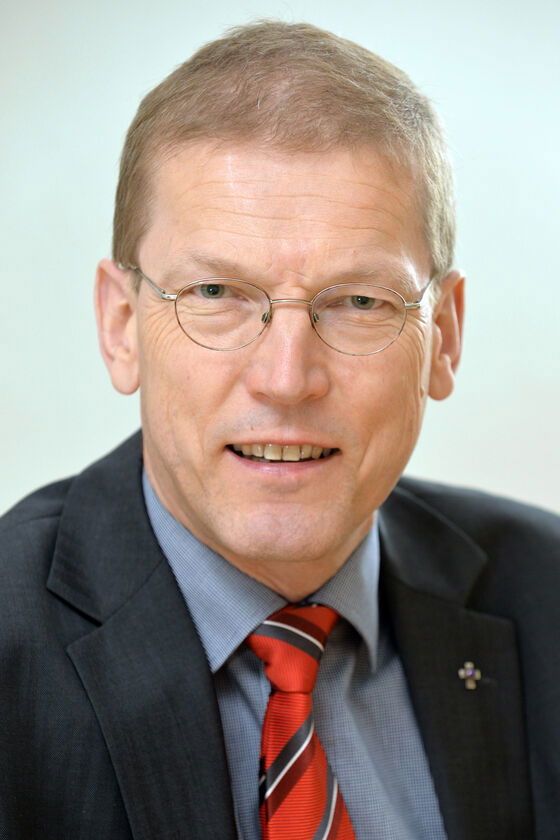 Ein Mann mit Brille und braunen Haaren, er trägt Anzug, Hemd und eine rote Krawatte mit blauen Streifen.