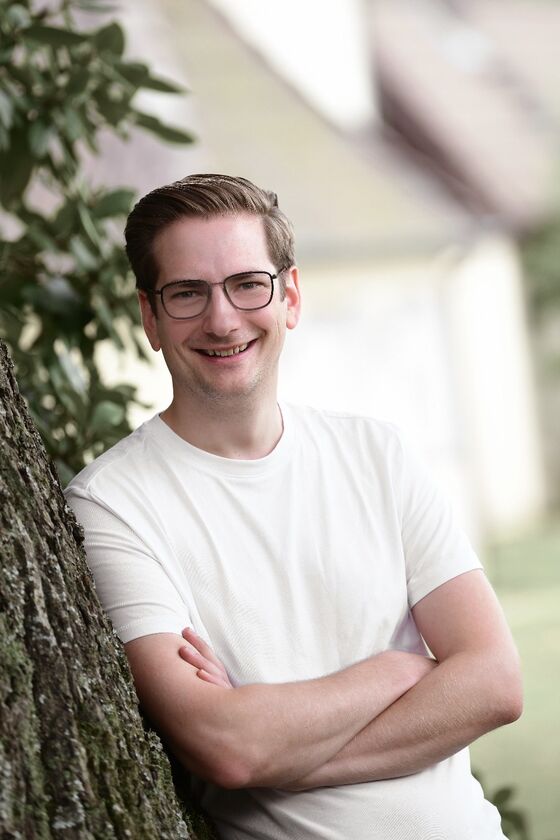 Eine männlich gelesene Person in weißem Shirt lehnt an einem Baum.