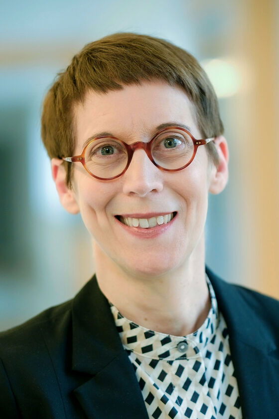 Eine weiblich gelesene Person mit dunklen kurzen Haaren, einer Brille mit dunkler Fassung und einem dunklen Jackett.