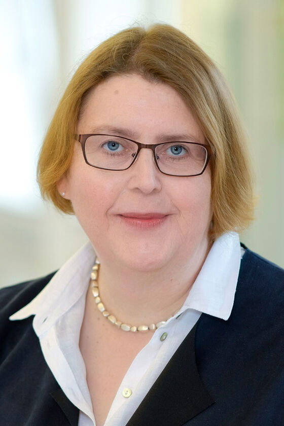 Das Bild zeigt eine weiblich lesbare Person mit mittelbraunem Pagenschnitt, einer Brille mit dunklem Rahmen, einer weißen Bluse und einem dunklen Jackett.