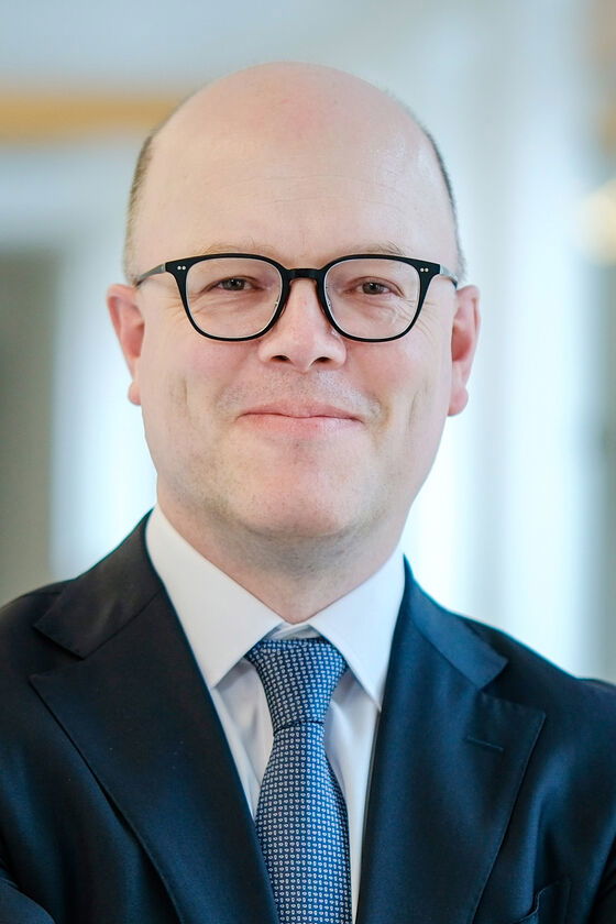 Eine männlich gelesene Person mit sehr kurzen Haaren, dunkler Brillenfassung, weißem Hemd, blauer Krawatte und dunklem Jackett.