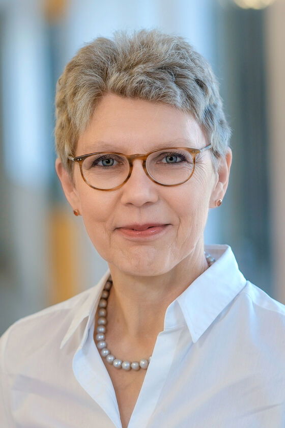 Das Bild zeigt eine weiblich lesbare Person mit kurzen grauen Haaren, Brille und weißer Bluse.