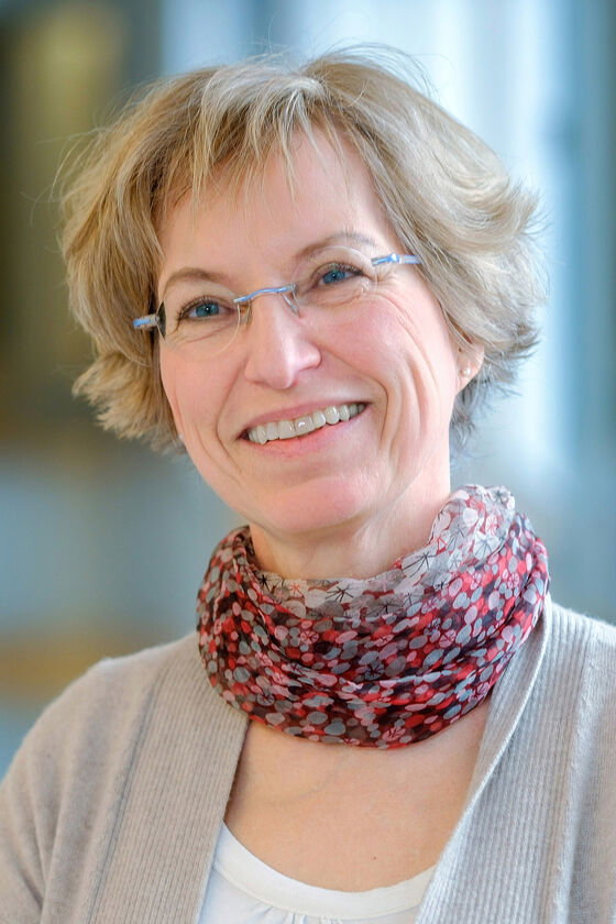 Das Bild zeigt eine weiblich lesbare Person mit kurzen blonden Haaren, einer Brille ohne Fassung, einem gemusterten Schal, einem weißen Oberteil und einer grauen Strickjacke darüber.