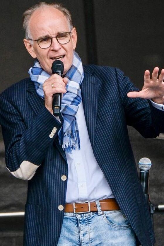 Das Bild zeigt eine männlich lesbare Person ohne Bart, mit kurzen grauen Haaren, einer Brille mit dunkler Fassung, einem hellblauen Schal, weißem Hemd und dunklem Sakko. Die Person hält ein Mikrophon in ihrer rechten Hand und hält die linke Hand auf Brusthöhe. 