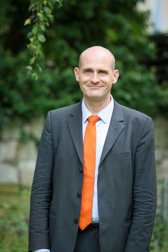 Ein Mann in grauem Anzug mit orangener Krawatte und Glatze steht im Grünen.