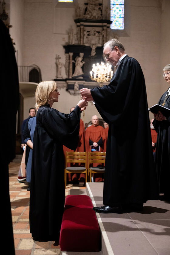 Ein älterer Mann im schwarzen Talar übergibt einer jüngeren Frau im schwarzen Talar ein Kreuz.