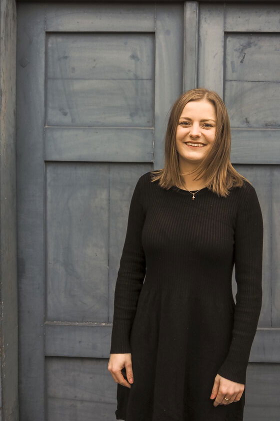 Junge Frau vor einer blauen Holztür. Sie trägt ein schwarzes Kleid und hat braune kinnlange Haare.