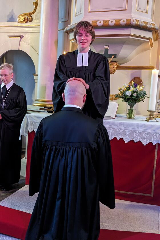 In einer hellen Kirche vor einem Altar mit roten Parament steht eine Pfarrerin, sie legt dem davor knienden Pfarrer die Hände segnend auf den Kopf. Links steht eine weitere Pfarrerin neben dem Altar.