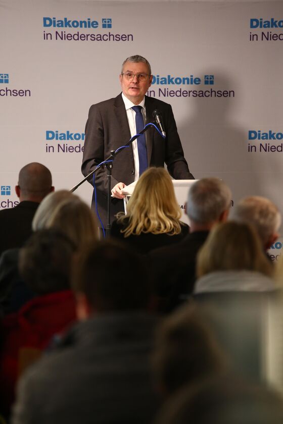 Eine männlich gelesene Person steht an einem Podium und hält eine Rede. Im Vordergrund sind die Rücken des Publikums zu sehen.