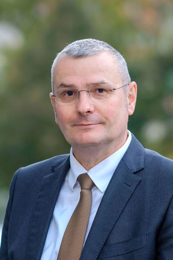 Eine männlich gelesene Person in blauem Jackett und mit Krawatte, mit raspelkurzen grauen Haaren und Brille, guckt aufmerksam in die Kamera.