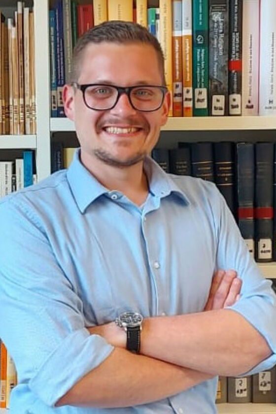 Eine als Mann lesbare Person mit Brille und hellblauem Hemd steht mit verschränkten Armen vor einer Bücherwand.