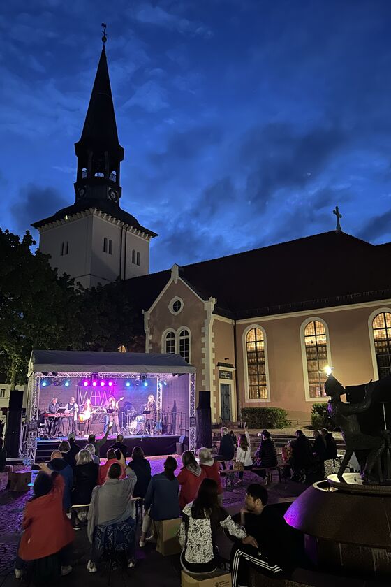 Gegen den blau-schwarzen Abendhimmel hebt sich eine Kirche mit spitzem Turm ab. Davor steht eine Bühne, in rot-lila-blaues Licht getaucht. Davor sitzen und stehen Menschen.