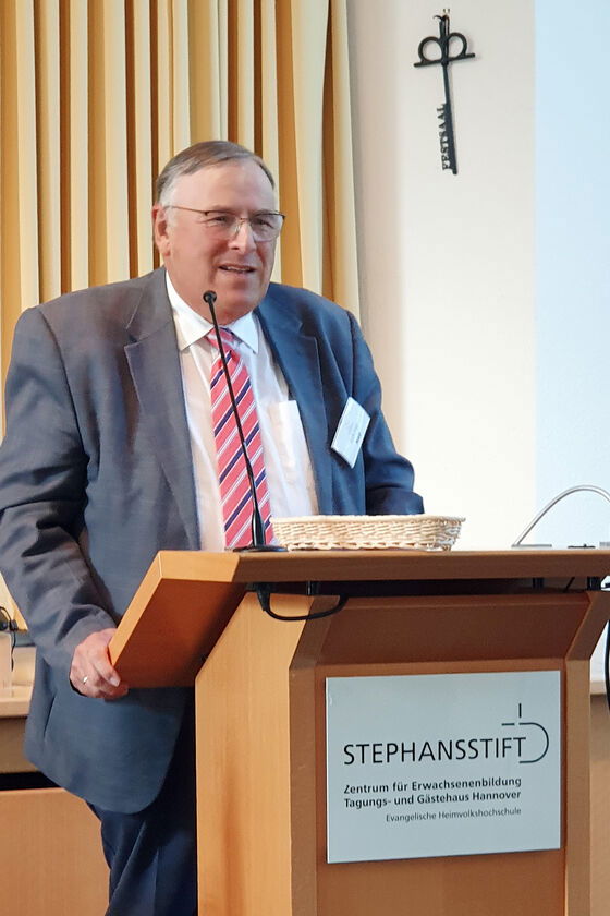 Eine ältere als Mann lesbare Person in Anzug und Krawatte spricht an einem Rednerpult.