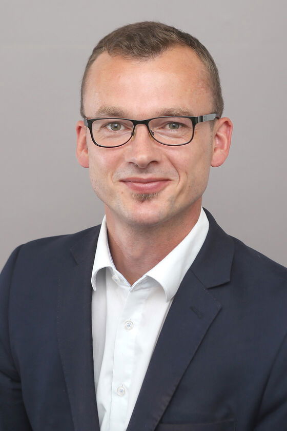 Eine als Mann lesbare Person mit kurzen Haaren und Brille. Er trägt ein weißes Hemd und ein dunkles Jacket.