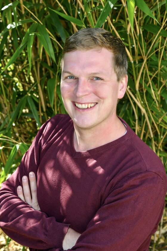 Eine männlich gelesene Person mit kurzen Haaren in rotem Shirt vor grünen Pflanzen.