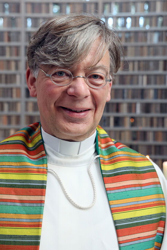 Eine als Mann lesbare Person mit grauen Haaren und Brille im weißen Talar mit bunter Stola.