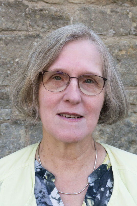 Eine weiblich gelesene Person mit halblangen grauen Haaren und einer Brille. Sie steht draußen vor einer Mauer. 