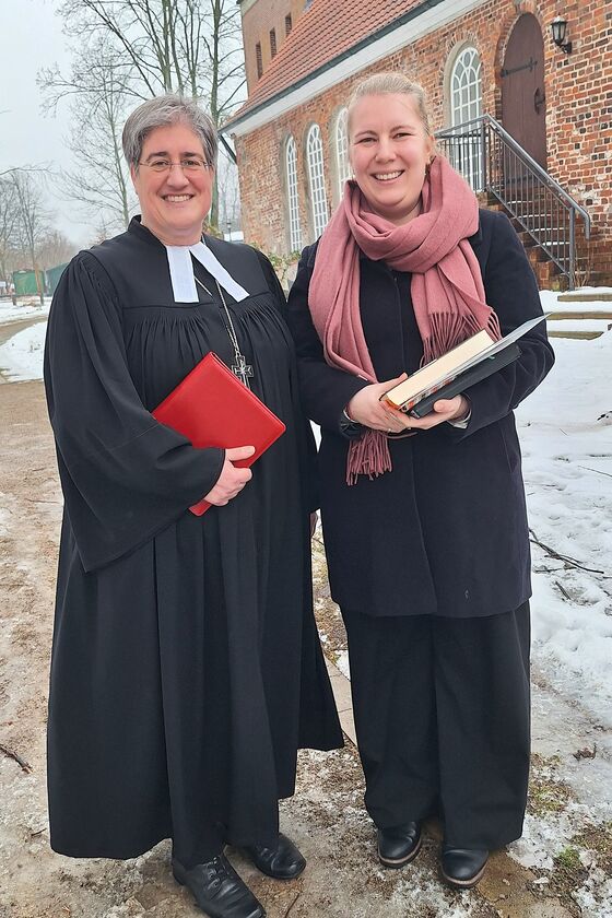 Zwei weiblich gelesene Personen, eine davon im Talar vor einer Kirche.