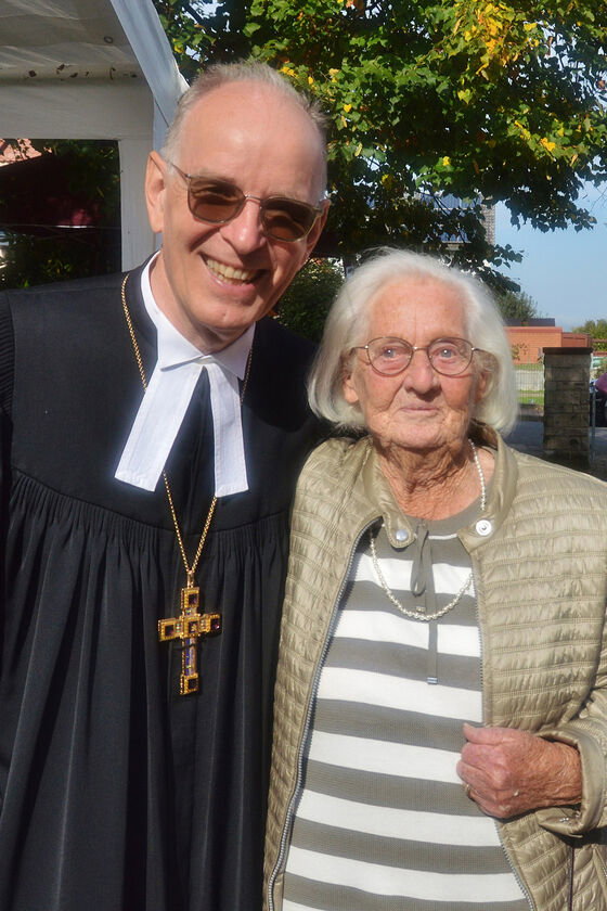 Eine als Mann lesbare Person im Talar und mit Bischofskreuz mit einer sehr alten Frau