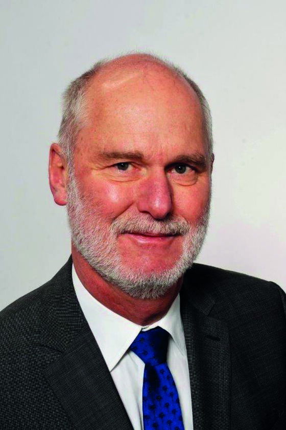 Eine männlich zu lesende Person im Portrait mit dunklem Anzug, weißem Hemd und blauer Krawatte