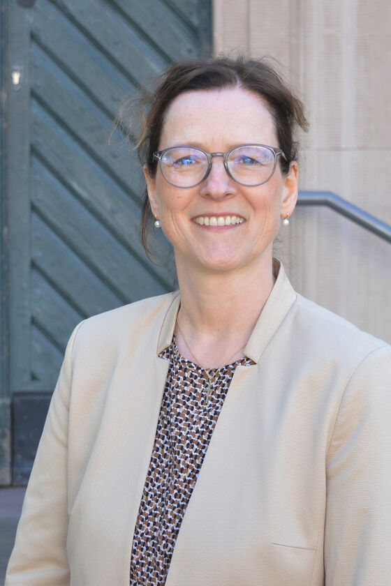 Eine als Frau lesbare Person mit Brille und hellem Jackett vor einer Kirchentür.
