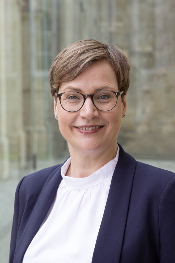 Eine weiblich lesbare Person mit kurzem braunen Haar, einer braunen Brille, weißem Oberteil und dunkelblauem Jackett.