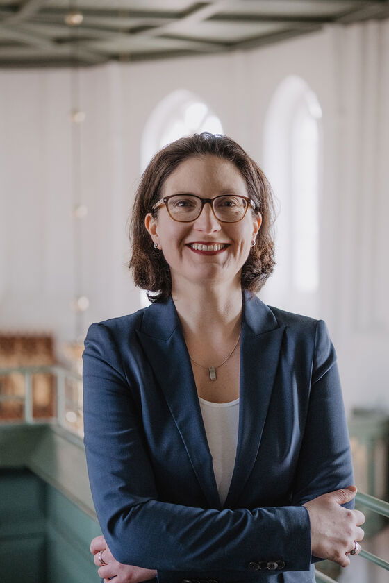 Eine weiblich zu lesende Person mit verschränkten Armen lächelt in die Kamera.