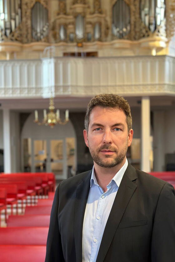 Eine mittelalte als Mann lesbare Person im hellblauen Hemd und dunklen Sakko steht in einer Kirche. Im Hintergrund ist eine Orgel zu sehen.
