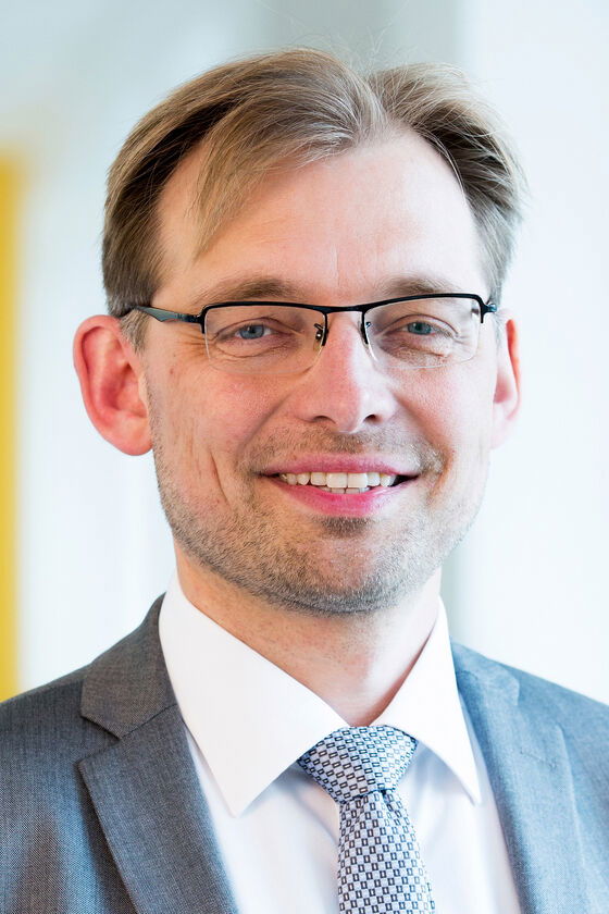 Ein Mann mit Brille mit grauem Anzug und Krawatte lächelt in die Kamera.