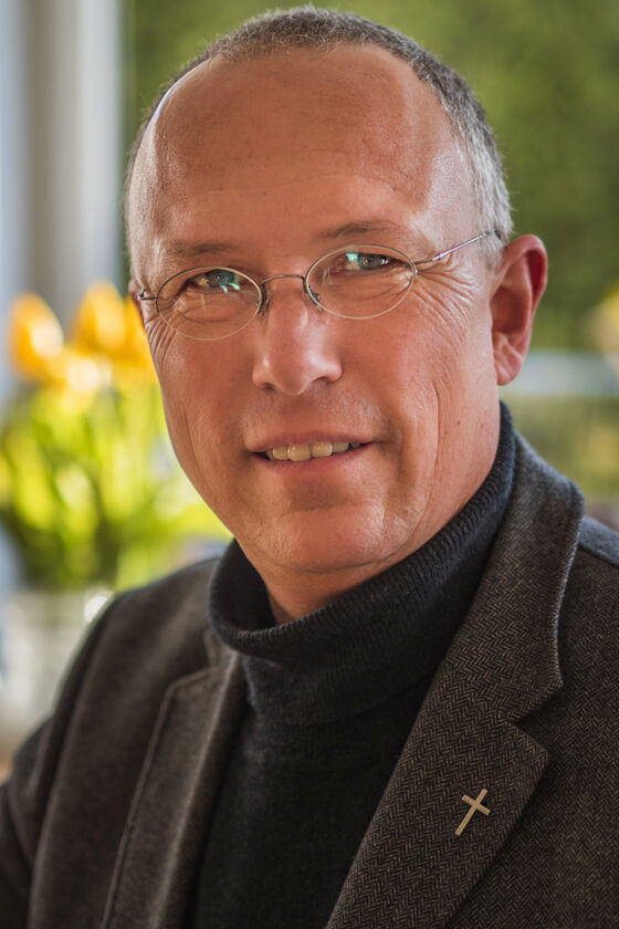 Ein Mann mit grauen kurzen Haaren und Brille. Er trägt einen schwarzen Pulli und ein graues Sakko mit einem kleinen Kreuz am Revers
