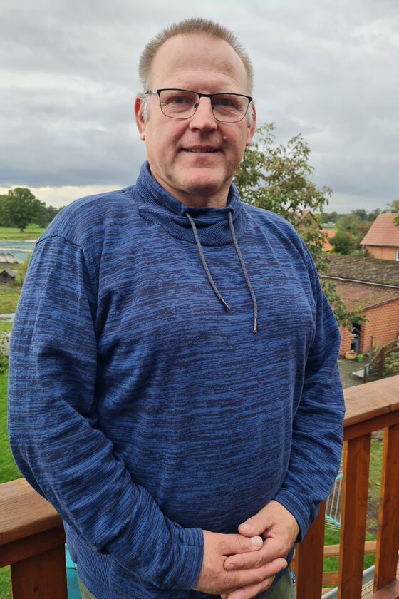 Ein Mann steht im Garten und schaut freundlich in die Kamera. Er trägt einen blauen Pulli, eine Brille und hat kurze graue Haare.