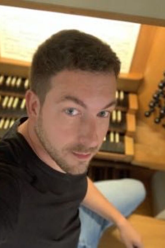 Ein junger Mann mit kurzen Haaren sitzt an einer Orgel und blickt über seine Schulter nach oben in die Kamera.