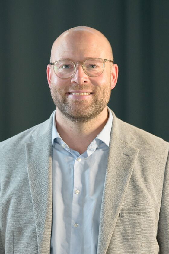 Ein Mann mit Glatze und Brille in beigem Sakko und hellblauem Hemd steht vor einem schwarzen Hintergrund.