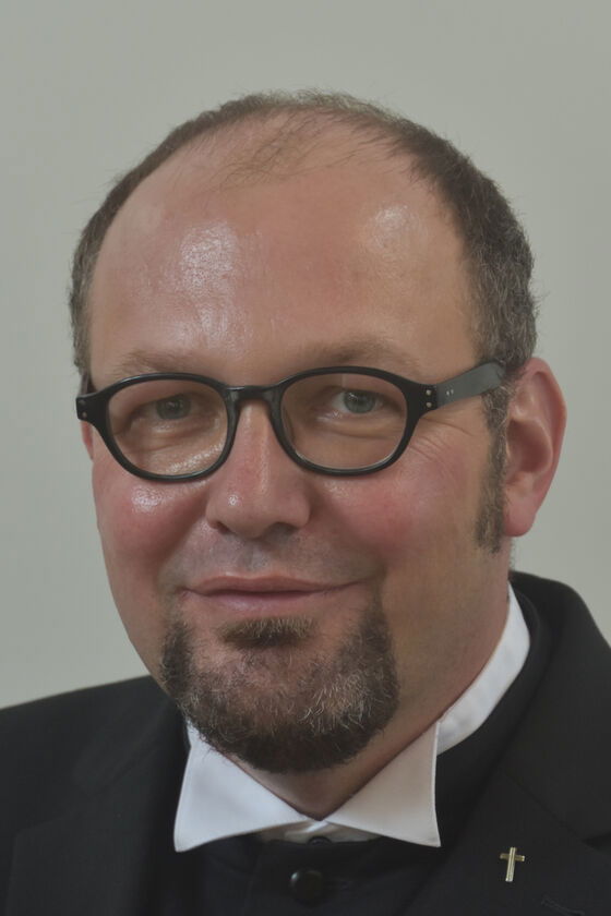 Ein Mann mit Halbglatze, kurzem Kinnbart und Brille lächelt verhalten.