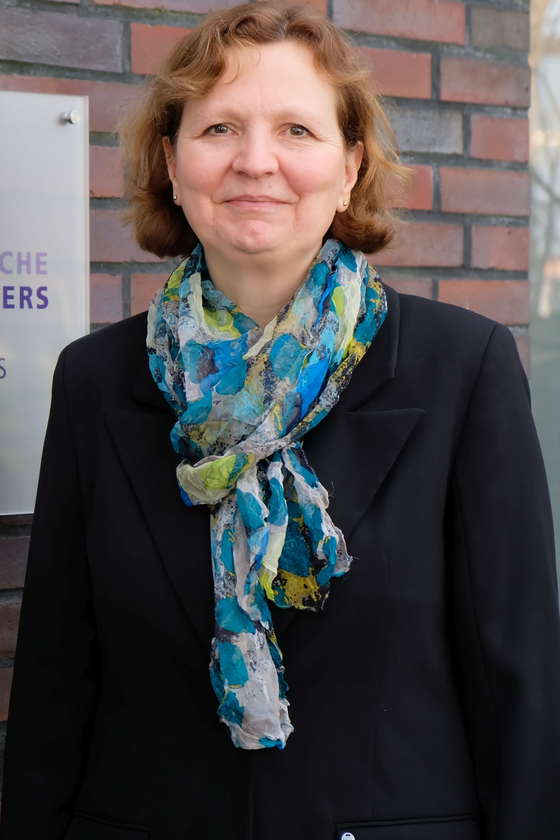 Eine Frau in schwarzem Pulli und mit blauem Schal steht vor einer Mauer. An der Mauer hängt ein Schild mit der Aufschrift: Landeskirche Hannovers.