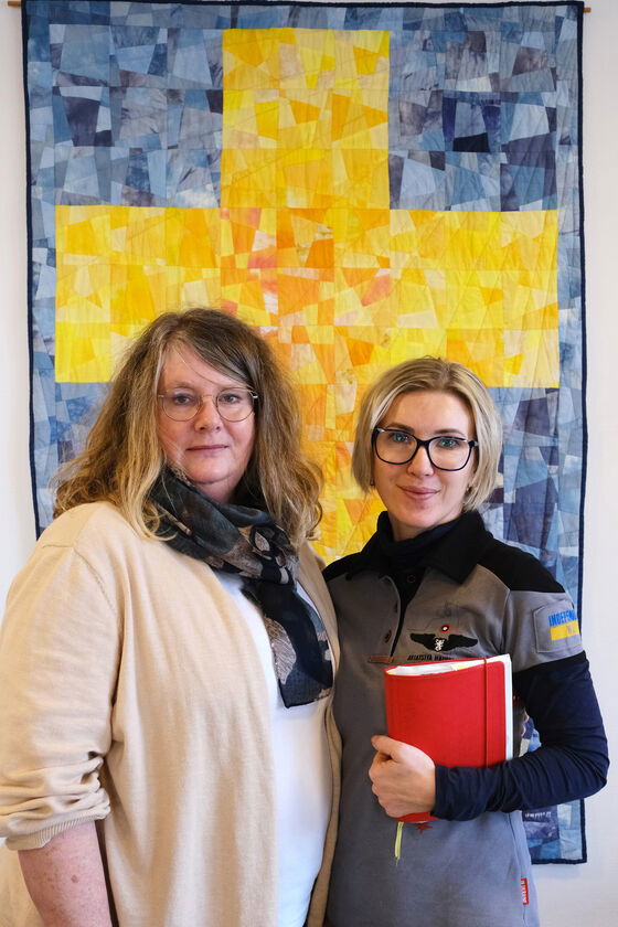 Zwei als Frauen lesbare vor einem Wandbehang mit einem gelben Kreuz auf blauem Grund.