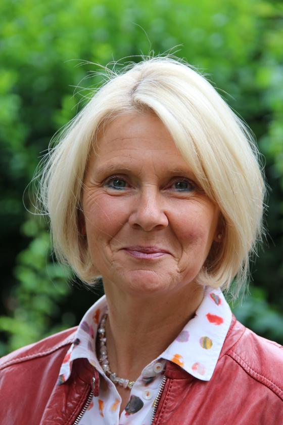 Porträt einer Frau mit kinnlangen blonden Haaren, einer roten Lederjacke und einer bunten Bluse.