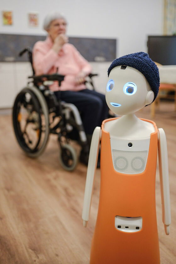 Eine weiblich gelesene Person sitzt im Rollstuhl. Vor ihr steht ein Roboter, der einem Jungen nachempfunden ist, Mund und Augen leuchten blau, mit orangener Latzhose und blauer Mütze.