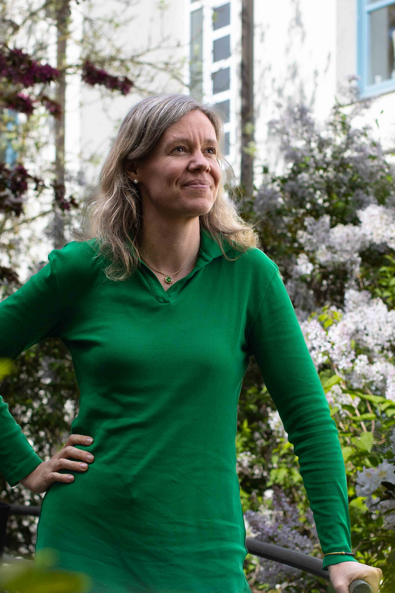 Eine weiblich gelesene Person in grünem Kleid steht an einer weiß blühenden Hecke.