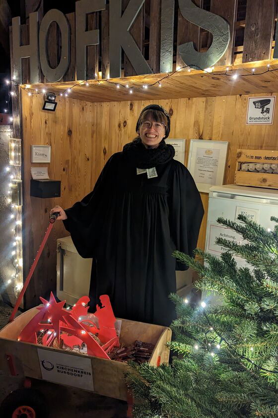 Eine weiblich gelesene Person in Winterjack und mit Mütze steht in einer Weihnachtsmarkt-Holzbude, vor sich eine Kiste und ein Tannenbaum.