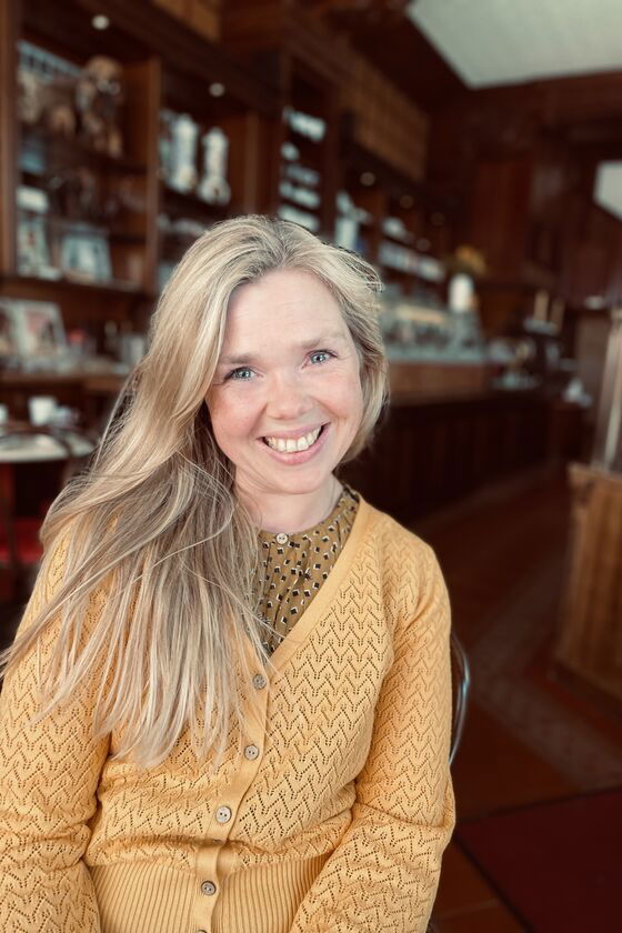 Eine blonde, weiblich gelesene Person in gelber Strickjacke sitzt vor einer großen, dunklen Holzwand und lächelt.