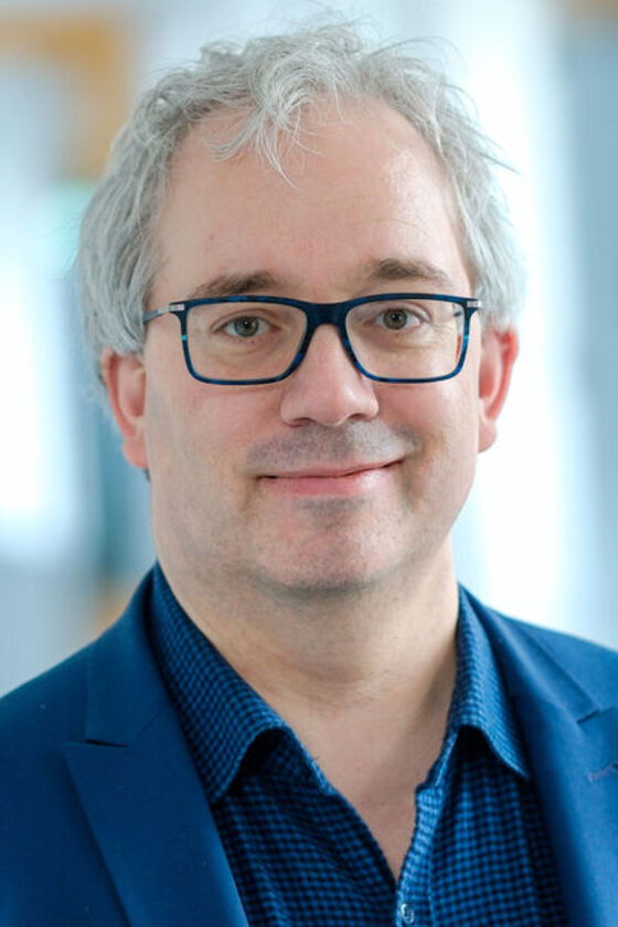 Eine als Mann lesbare Person mit Brille, blauem Hemd und blauem Jackett